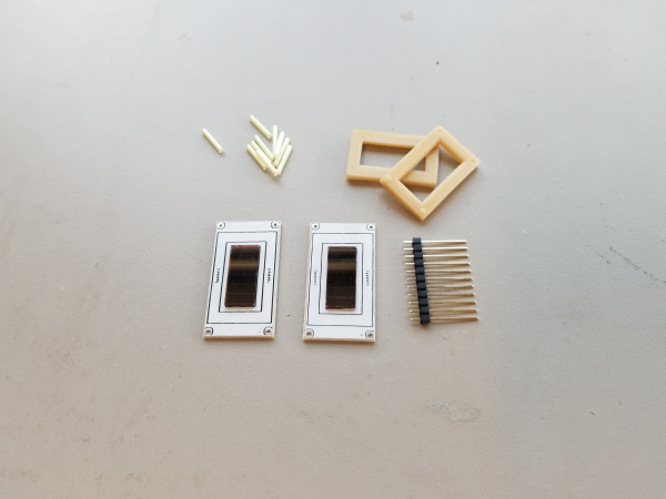 All of the parts of the kit laid out on a gray background. Two white PCBs with PV cells centered on each, a pair of beige rectangular silicone gaskets, a strip of long pin headers, and a small pile of tritium glow tubes.