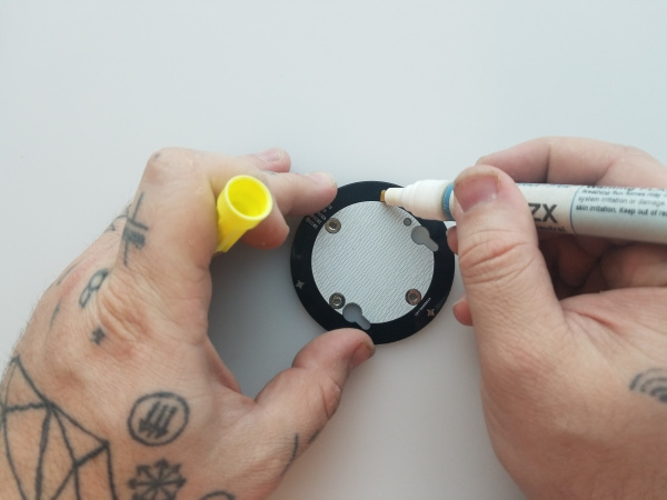 Close up of a hand holding a flux pen and applying flux to one of the small solder pads on the base board in preparation for soldering the pogo pin.