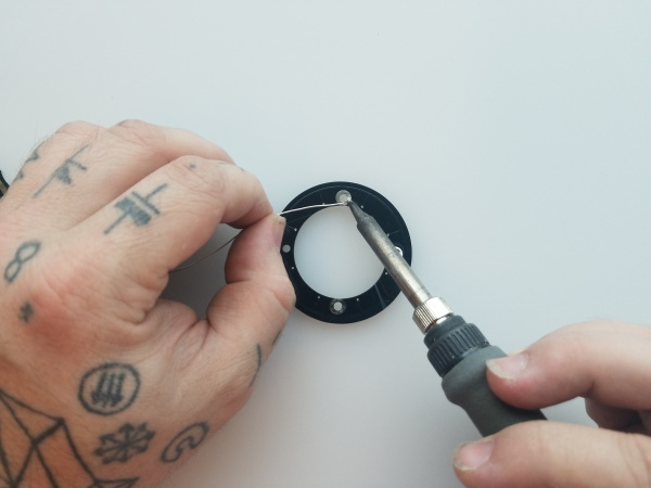 Hands hold a soldering iron and a length of solder against the exposed copper surrounding a standoff hole in the base board