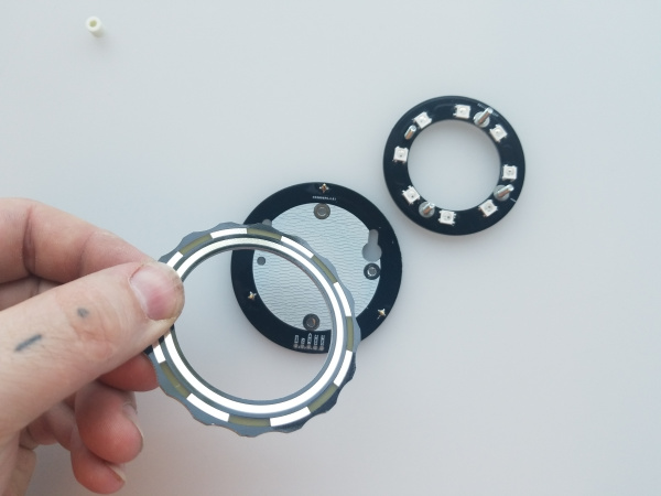 A ring shaped board is laying face up to the right with LEDs on its face and bolts protruding toward the camera. In the center of the photo another round board is sitting with pogo pins facing up. A hand holds another ring shaped board overtop of the round board.