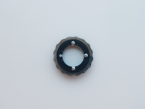 top down view of the completed assembly. A round contraption with a notched ring along the outside, trapped in place by a top retaining ring. You can see through the center of the retaining ring to the base board below which is covered in white silkscreen. This is designed to reflect the LEDs which are mounted on the underside of the retaining ring.