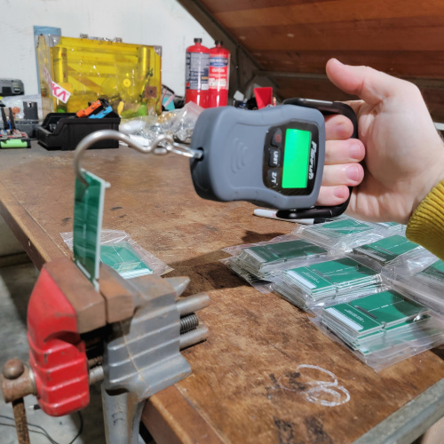 One of the small PCBs is clamped in a bench vise on the workbench and a hand holds the fishing scale with the hook connected through a hole on the PCB, preparing to pull on the scale and break teh board.