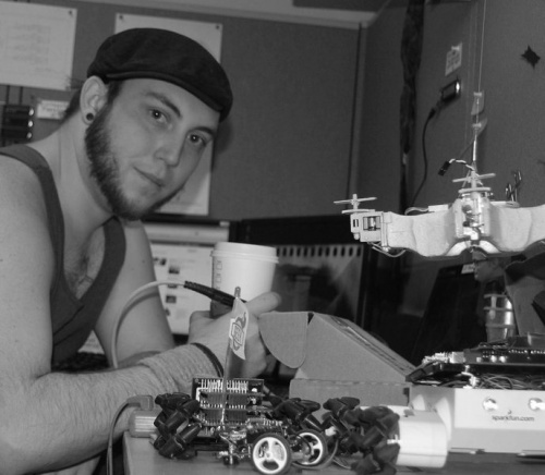 black and white, low-res photo of me sitting at a desk with several small robots on the desk in front of me. I'm holding a soldering iron and a cup of coffee. I'm wearing a tank top and a flat cap.