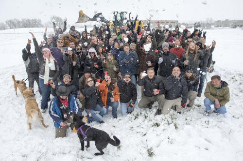 Group photo of people gathered in front of an excavator holding small plastic shovels. There's snow on the ground and in the air.