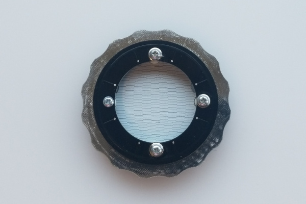 top down view of the completed assembly. A round contraption with a notched ring along the outside, trapped in place by a top retaining ring. You can see through the center of the retaining ring to the base board below which is covered in white silkscreen. This is designed to reflect the LEDs which are mounted on the underside of the retaining ring.